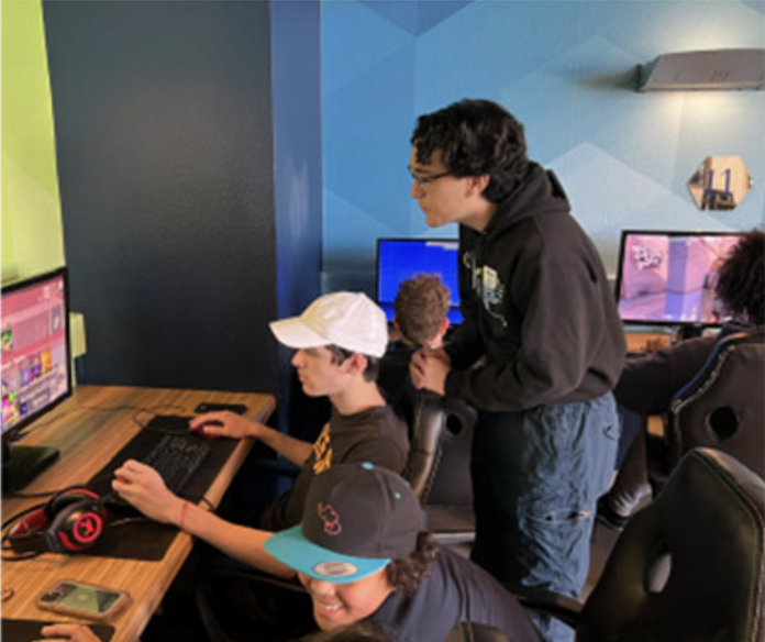 Three people are gathered around a computer desk. Two are seated and gaming, while one stands and observes.